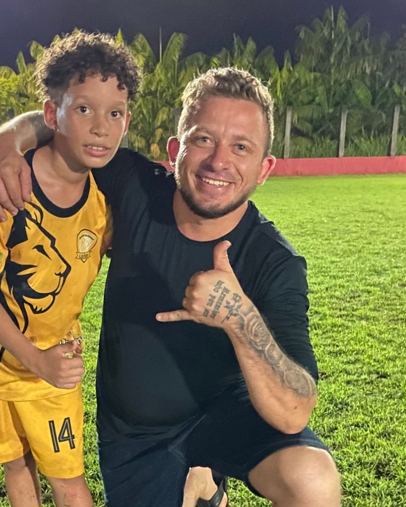 Marlon Rosa ajoelhado no gramado, à noite, abraçado ao filho, que veste o uniforme amarelo da escolinha