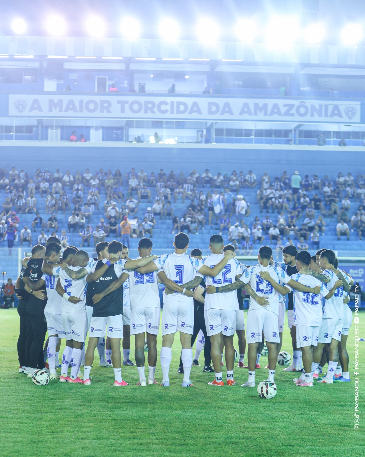 Jogadores do Paysandu abraçados em roda no gramado antes do jogo, com a torcida e a faixa "A maior torcida da Amazônia" ao fundo